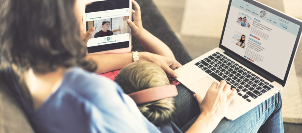 A parent with a laptop on their lap browsing Gander's blog, a child wearing headphones is also on their lap watching a video on an ipad