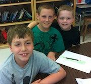 Three young boys at a table with homework smiling